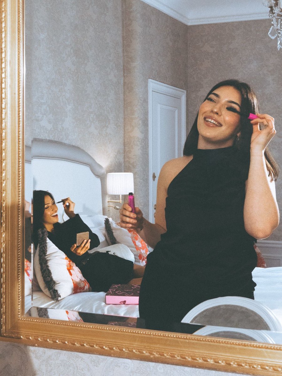 Une femme se maquille devant un grand miroir, son amie allongée sur le lit derrière elle.